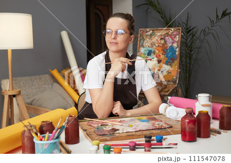 Craft education studio. Professional artist workshop. Artwork inspiration session. Pensive Caucasian woman painter wearing brown apron in art studio drawing picture Craft education studio. Professional artist workshop. Artwork inspiration session. Pensive Caucasian woman painter wearing brown apron in art studio drawing picture 111547078