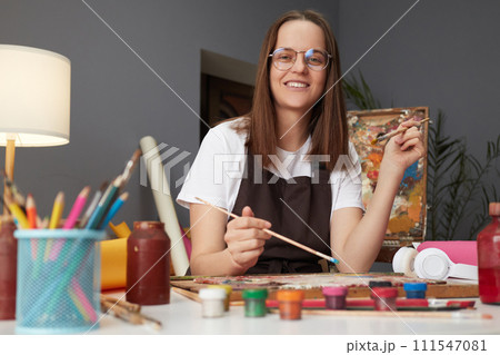 Artist at work in the studio. Creative painting and drawing. Artistic hobby and passion. Caucasian woman painter wearing brown apron in art studio drawing picture Artist at work in the studio. Creative painting and drawing. Artistic hobby and passion. Caucasian woman painter wearing brown apron in art studio drawing picture 111547081