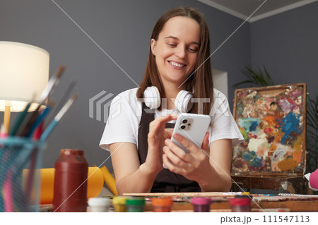 Cheerful joyful smiling Caucasian woman wearing brown apron painting in art creative studio and using smartphone chatting with friends while having break 111547113