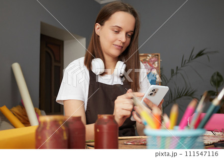Concentrated Young Caucasian woman wearing brown apron painting with brush in artist studio and using cell phone looking at mobile phone reading messages 111547116