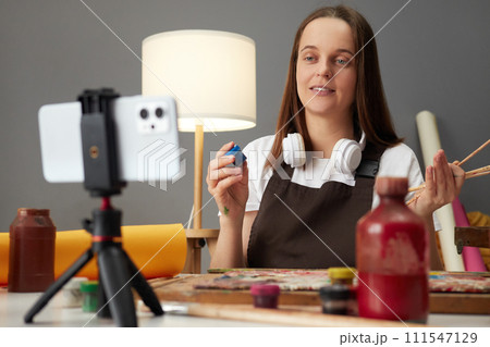 Brown haired woman wearing brown apron painting using mobile phone on tripod at her workplace broadcasting online master class teaching followers to use contemporary technics Brown haired woman wearing brown apron painting using mobile phone on tripod at her workplace broadcasting online master class teaching followers to use contemporary technics 111547129