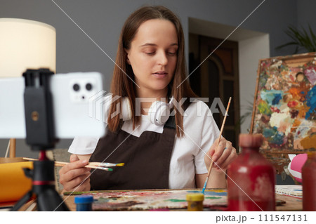 Busy concentrated young Caucasian woman wearing brown apron painting with brush in artist studio using smartphone and tripod drawing online and explaining technic to students 111547131