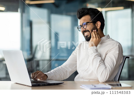 Toothache in a young Indian male businessman. Sitting at the workplace in the office at the table and holding a hand to the cheek, doing a massage, suffering from inflammation. 111548383