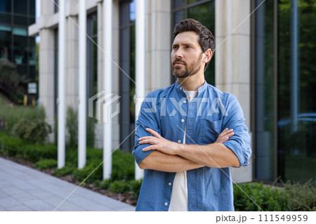 Mature bearded businessman in casual clothing confidently standing with arms crossed outside a modern office; portrays a relaxed professional vibe. 111549959