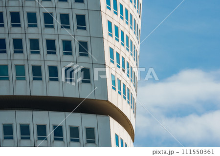 Closeup of a modern curved building with a pattern of white panels and bluetinted windows, accented by a brown horizontal band, against a partly cloudy sky, likely in Malmo. 111550361