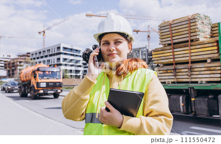 Young architect woman in white hardhat and safety vest with digital tablet talking on smartphone near modern building. Female engineer. Logistics, shipping and construction worker on the phone Young architect woman in white hardhat and safety vest with digital tablet talking on smartphone near modern building. Female engineer. Logistics, shipping and construction worker on the phone 111550472