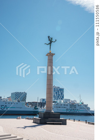 A statue with outstretched wings atop a column overlooks a promenade by the sea, with a FORSEA ferry and modern checkered buildings, likely in Helsingborg. A statue with outstretched wings atop a column overlooks a promenade by the sea, with a FORSEA ferry and modern checkered buildings, likely in Helsingborg. 111550586