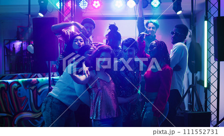 Men and women taking photos at party and dancing together under stage lights. Young adults having fun with pictures on smartphone, enjoying disco event at social gathering. Handheld shot. 111552731