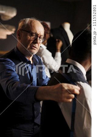Professional expert suitmaker checking custom made suit fitting assisted by couturier. Apprentice tailor helping master fashion designer with sartorial measurements for upcoming collection, close up 111552801