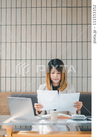 Freelancer concept, Focused businesswoman in a beige blazer reviews paperwork with her laptop at a cafe table, embodying professional multitasking. Freelancer concept, Focused businesswoman in a beige blazer reviews paperwork with her laptop at a cafe table, embodying professional multitasking. 111553352