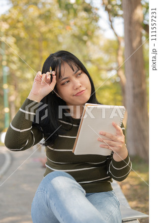 A woman is sitting on a bench in a green park with her notebook, contemplating ideas for inspiration 111557231