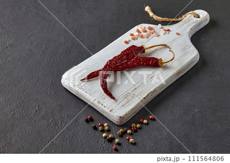 two red hot peppers, scattered peppercorns and pink salt on a white wooden cutting board on a black stone background two red hot peppers, scattered peppercorns and pink salt on a white wooden cutting board on a black stone background 111564806