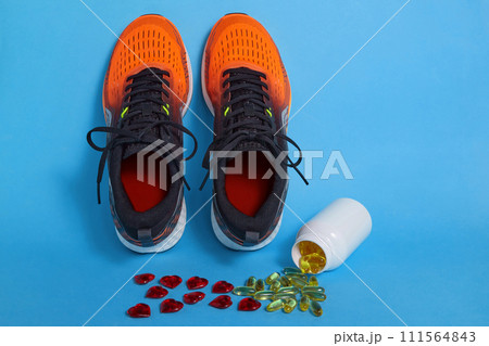 Scattered yellow vitamin D capsules from a jar and red decorative hearts and orange running sneakers on a blue background 111564843