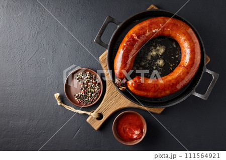 Fried pork sausage on a black cast iron pan on a wooden board and peppercorns with tomato sauce on a black stone table. Top view with copy space Fried pork sausage on a black cast iron pan on a wooden board and peppercorns with tomato sauce on a black stone table. Top view with copy space 111564921