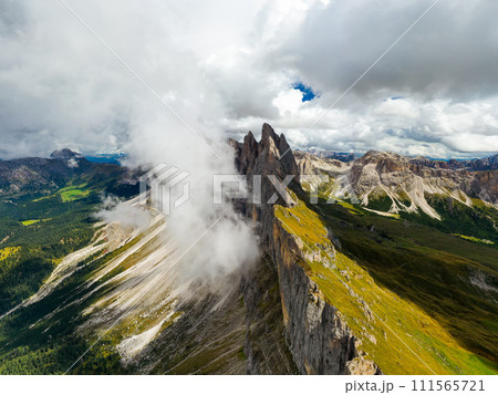 White cloud surrounds giant Seceda ridgeline in Italian Alps. Rocky mountain range with steep slopes under cumulus clouds aerial view 111565721