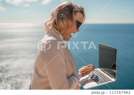 Freelance women sea working on the computer. Good looking middle aged woman typing on a laptop keyboard outdoors with a beautiful sea view. The concept of remote work. Freelance women sea working on the computer. Good looking middle aged woman typing on a laptop keyboard outdoors with a beautiful sea view. The concept of remote work. 111567962