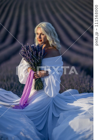 Blonde woman poses in lavender field at sunset. Happy woman in white dress holds lavender bouquet. Aromatherapy concept, lavender oil, photo session in lavender 111568000
