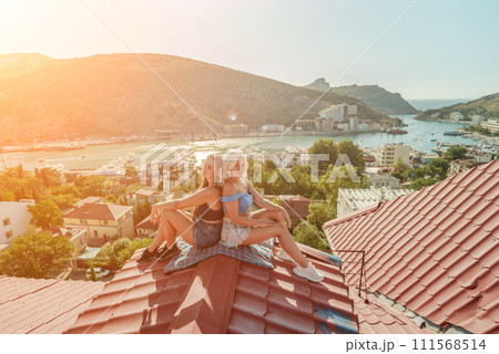 Two women sitting on a red roof, enjoying the view of the town and the sea. Rooftop vantage point. In the background, there are several boats visible on the water, adding to the picturesque scene. 111568514