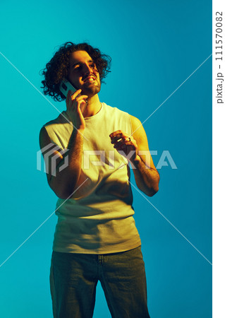 Young man with curly hair and unshaved face, in casual clothes talking on mobile phone against blue background in neon light 111570082
