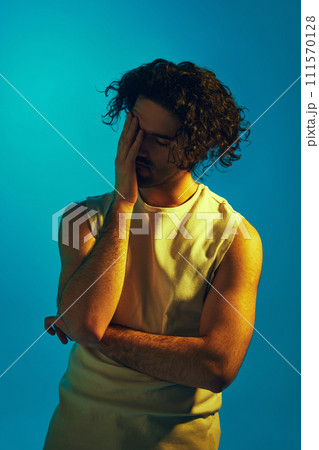 Man in tank top standing with eyes closed, hand on forehead against blue background in neon light. Feeling stressful and annoyed 111570128