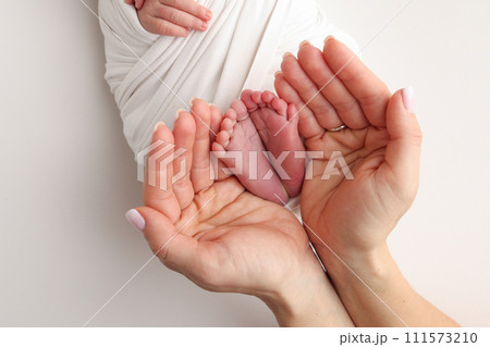 The palms of the father, the mother are holding the foot of the newborn baby in a white blanket. Feet of the newborn on the palms of the parents. Studio macro photo of a child's toes, heels and feet. 111573210