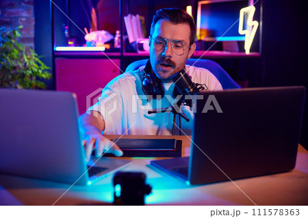 Man in vibrant studio setting speaking into microphone and typing on laptop keyboard in neon light at home-studio. 111578363