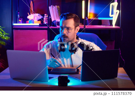 Focused man in headphones and glasses podcasting and looking to laptop, surrounded by neon lights at home-studio. 111578370
