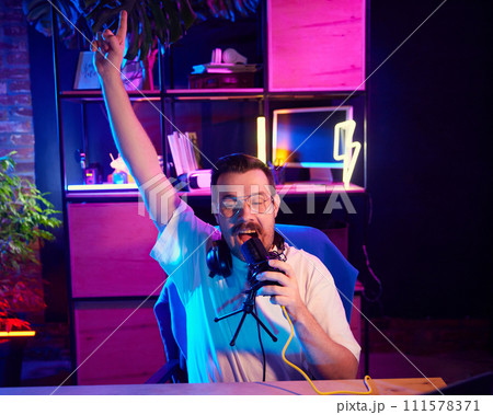 Portrait of happy and excited broadcaster, blogger recording audio in colorful, neon-lit workspace, at home-studio. Portrait of happy and excited broadcaster, blogger recording audio in colorful, neon-lit workspace, at home-studio. 111578371