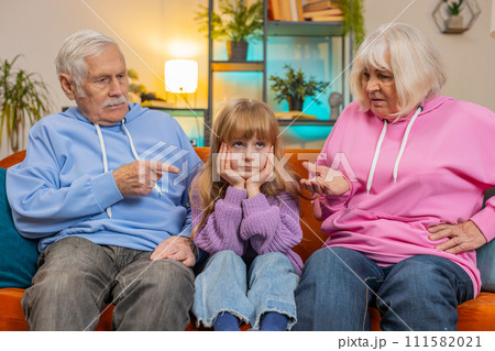 Irritated grandfather and grandmother scolding granddaughter child kid for bad behavior on home sofa Irritated grandfather and grandmother scolding granddaughter child kid for bad behavior on home sofa 111582021