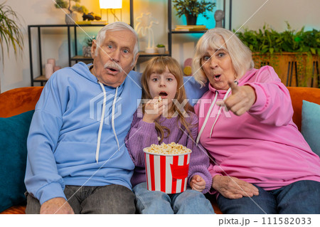 Child girl granddaughter grandfather grandmother eating popcorn watching movie film on home sofa 111582033