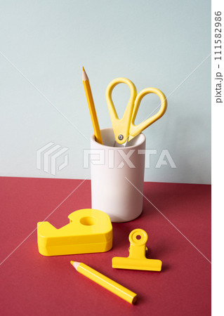 Office supply stationery. Yellow colored pencil, scissors, tape dispenser, clip on red desk. gray wall background. workspace 111582986