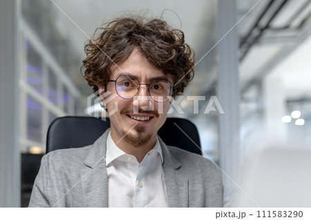 Smiling young businessman with curly hair and glasses working at his computer in a contemporary office setting. 111583290