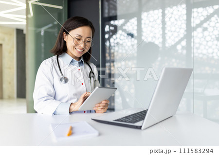 Young smiling Asian woman doctor, student sitting in the hospital office at the work table in a white coat, working on studying on a laptop, making notes in a notebook. 111583294