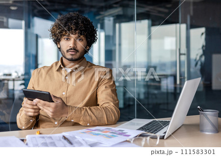 Portrait of a serious and worried young Indian man sitting in the office at a desk, working with documents and at a laptop, holding a calculator in his hands, having problems with calculations. 111583301