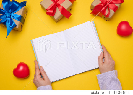 An open notebook with empty white sheets and gift boxes around on a yellow background, top view. Festive background for wish An open notebook with empty white sheets and gift boxes around on a yellow background, top view. Festive background for wish 111586619