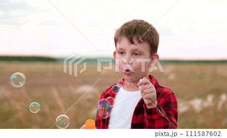Boy spends day blowing soap bubbles in cultivated field. Boy child stands in nature savouring simple pleasure of soap bubbles. Boy in checkered shirt playing with soap bubbles stand in field Boy spends day blowing soap bubbles in cultivated field. Boy child stands in nature savouring simple pleasure of soap bubbles. Boy in checkered shirt playing with soap bubbles stand in field 111587602