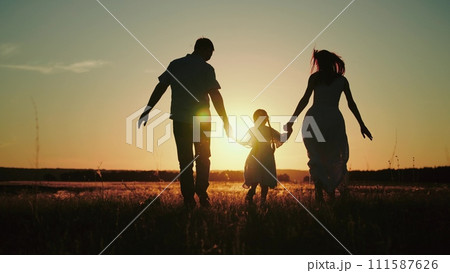 Small girl happily runs hand in hand with parents on field to release energy after day at kindergarten. Small girl loves to run across field at sunset having fun with papa and momma at sunset Small girl happily runs hand in hand with parents on field to release energy after day at kindergarten. Small girl loves to run across field at sunset having fun with papa and momma at sunset 111587626