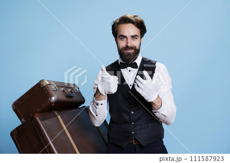 Positive bellboy on smartphone app, enjoying excellent news on online website in studio. Confident happy hotel porter in suit and tie finds out great information for tourism industry. 111587923