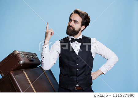 Male bellhop worker getting an idea, finding solution in front of camera next to trolley bags. Hotel porter in formal clothes being genius and thinking about something to solve problems. Male bellhop worker getting an idea, finding solution in front of camera next to trolley bags. Hotel porter in formal clothes being genius and thinking about something to solve problems. 111587990