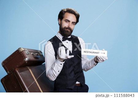 Hotel worker holds restaurant sign to help guide tourists with directions, posing with area pointer on camera. Young classy bellboy shows indication symbol for the dining room. 111588043