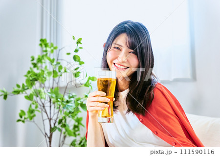 休日の明るい時間から自宅の部屋でビールを手に持ち1人で飲む20代から30代のカメラ目線の笑顔の女性 111590116