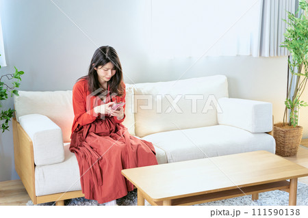 部屋のソファに座って困って悩んだ表情でスマートフォンを操作する30代の1人の女性 111590138