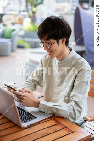 A happy Asian man is using his smartphone while working remotely at a coffee shop. A happy Asian man is using his smartphone while working remotely at a coffee shop. 111590718