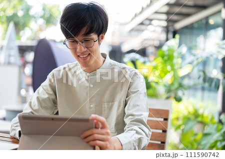 A happy young Asian man is working remotely at a coffee shop in the city, using his digital tablet. 111590742
