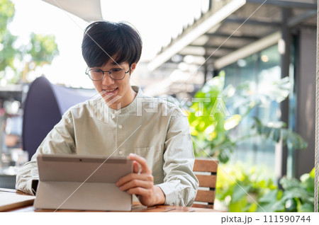 A happy young Asian man is working remotely at a coffee shop in the city, using his digital tablet. A happy young Asian man is working remotely at a coffee shop in the city, using his digital tablet. 111590744