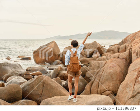 Tropical Sunset Adventure: A Happy Woman Walking on a Beach with a Backpack, Enjoying a Carefree Trip and Smiling at the Ocean Tropical Sunset Adventure: A Happy Woman Walking on a Beach with a Backpack, Enjoying a Carefree Trip and Smiling at the Ocean 111596837