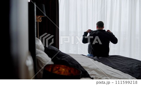 An adult man in a shirt in a room looks out the window. Businessman morning. A cozy room with a bed on which the guy sits with his back. A guy in a black shirt is sitting on the bed. An adult man in a shirt in a room looks out the window. Businessman morning. A cozy room with a bed on which the guy sits with his back. A guy in a black shirt is sitting on the bed. 111599218