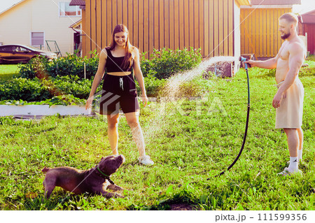 Shirtless white man waters dog with water from garden hose, young woman in her 20s smiles as she watches her playful pet. Shirtless white man waters dog with water from garden hose, young woman in her 20s smiles as she watches her playful pet. 111599356