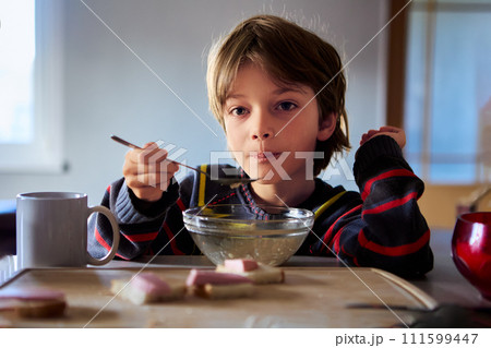 Unhappy male child sits in kitchen table with bowl of cereal in dimly lit kitchen. 111599447