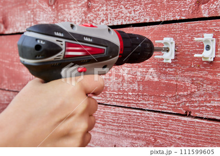Using cordless screwdriver to install external pipeline holder latches on weathered red wood wall. 111599605